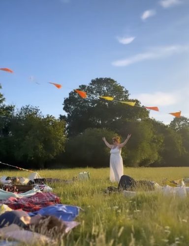 Debs Chakra Meditation A person in a white dress stands in a field, surrounded by colourful fabric and bunting.