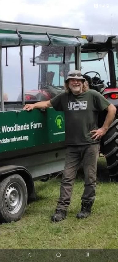 Meet Alex, Volunteer A person standing beside a green trailer and a tractor on a farm.