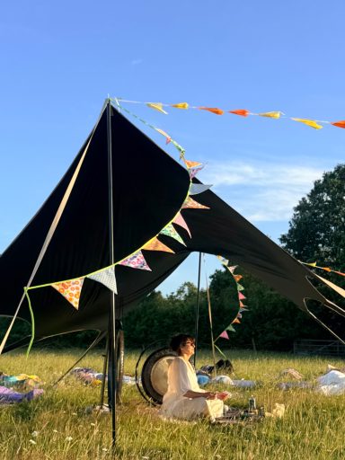 Gong with Dawn Person sitting beneath a canopy with colourful bunting and gongs in a grassy field.