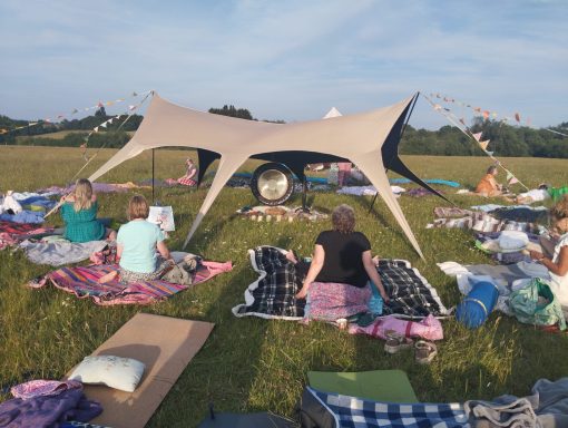 Gathering around the Gong People sitting on blankets under a large tent in a grassy field, enjoying a sunny day.