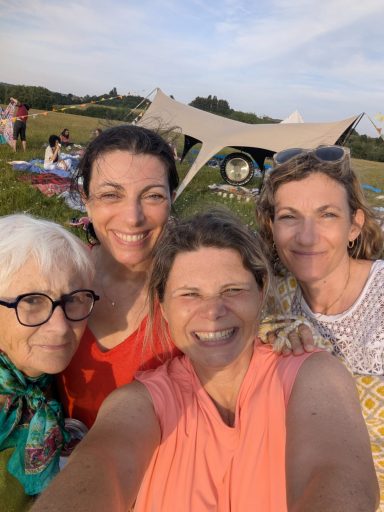 Happy Community Four women smiling together outdoors in a grassy area, with tents and people in the background.